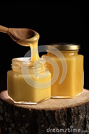 On a wooden log there are two glass jars with honey on a dark background. take honey out of the jar with a wooden spoon Stock Photo