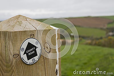 Wooden post with Devon County Road sign and green fields beyond. Stock Photo