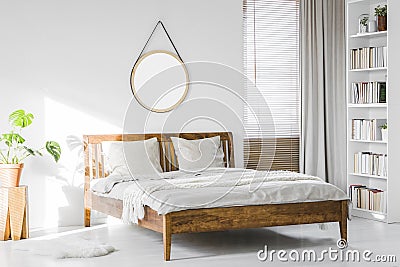 A wooden rustic bed frame and a home library bookcase in a natural, sunlit hotel room interior with white walls Stock Photo