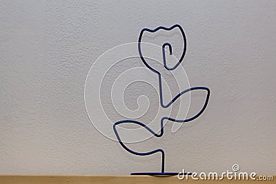 On a wooden shelf, against a background of white wallpaper, stands a figure of a rose flower made of steel wire. Stock Photo