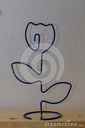 On a wooden shelf, against a background of white wallpaper, stands a figure of a rose flower made of steel wire. Stock Photo