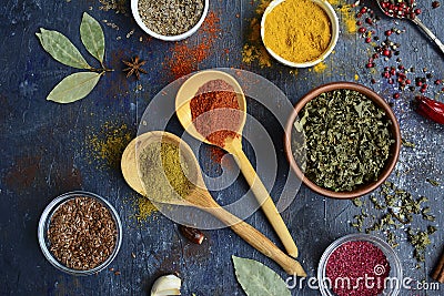 Wooden spoons with yellow and orange spices, different cups with dried herbs, garlic, bay leaf on a dark blue background Stock Photo