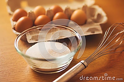 On a wooden table are the ingredients: chicken eggs in a package and sugar in a glass bowl, and next to it is a whisk. Cooking Stock Photo