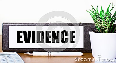 On a wooden table there is a folder for documents with the text EVIDENCE, a green plant in a pot, a pen and a calculator. Business Stock Photo