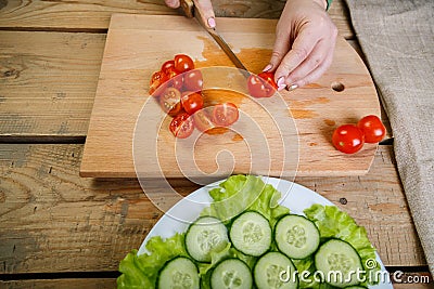 On a wooden table, a woman knifes a regimen of cherry tomatoes on slices for vegetable salad Stock Photo