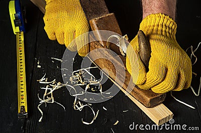 Woodworker using a hand plane to clean up a wooden board. Hands of the master in gloves close up at work. Working environment in Stock Photo