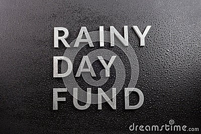 The words rainy day fund laid with silver metal letters on black background, covered with water drops, flat lay perspective, Stock Photo