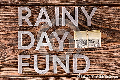 Words RAINY DAY FUND laid on wooden surface by metal letters with small roll of us dollar banknotes, flat lay from above Stock Photo