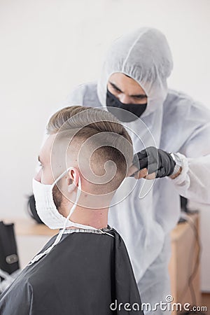 The work of the barber during the coronavirus, the hairdresser trim the client in a mask and a protective suit, quarantine Stock Photo