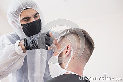 The work of the barber during the coronavirus, the hairdresser trim the client in a mask and a protective suit, quarantine Stock Photo