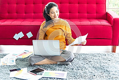 Work from home concept. Young Asian beautiful woman using laptop with gadget while sitting on gray carpet at home Stock Photo