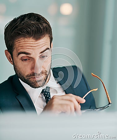 The work just keeps piling up. a tired young businessman holding a pair of glasses while working on a computer at the Stock Photo