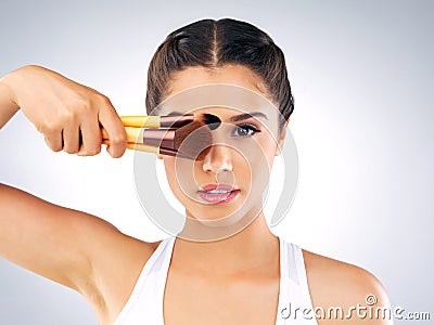 They work just like magic. Studio portrait of a beautiful young woman holding up makeup brushes against her face against Stock Photo