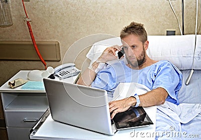 Workaholic business man in hospital room lying in bed sick and injured working with mobile phone computer laptop Stock Photo