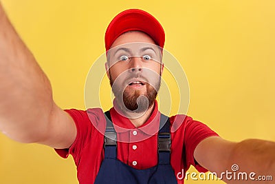 Worker man with wearing red cap and blue uniform taking selfie, looking at camera with big eyes POV. Stock Photo