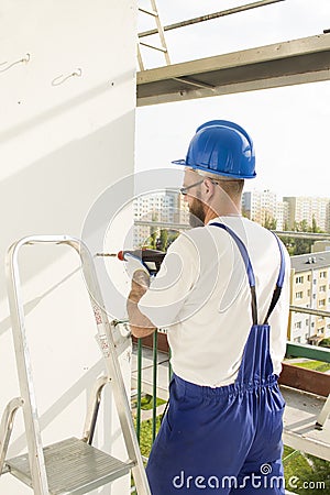 Worker in work attire, protective gloves and a helmet on site. Drilling the hole with a drill in the wall. Stock Photo