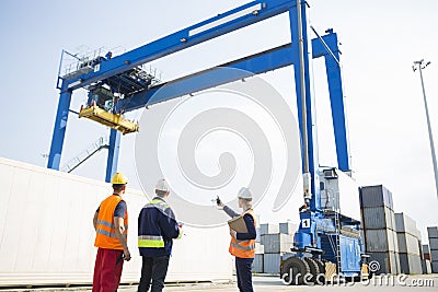 Workers discussing against large crane loading container at shipping yard Stock Photo