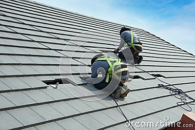 Workers are installing gray roof tiles wearing seat belts to ensure safe working at heights Stock Photo
