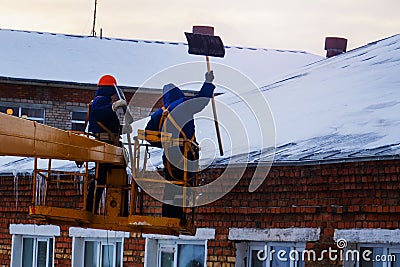 Workers in overalls and orange helmets on the crane basket remove icicles from roof of the house on a winter day - cleaning the Editorial Stock Photo