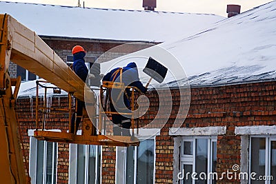 Workers in overalls and orange helmets on the crane basket remove icicles from roof of the house on a winter day - cleaning the Editorial Stock Photo
