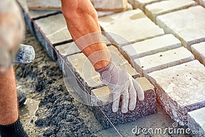 A workman`s gloved hands use a hammer to place stone pavers. Worker creating pavement using cobblestone blocks and granite stones. Stock Photo