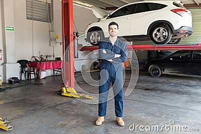 Workman Wearing Coveralls While Crossing Arms Against Car Lift Stock Photo