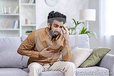 Worried young man with hand on chest feeling chest pain, expressing stress and anxiety in home setting Stock Photo