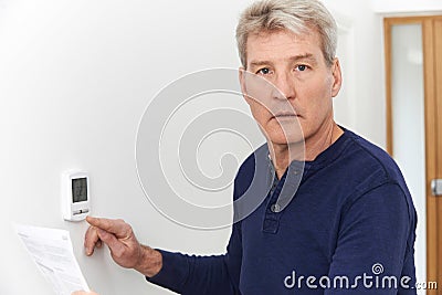 Worried Mature Man With Bill Turning Down Central Heating Thermostat Stock Photo