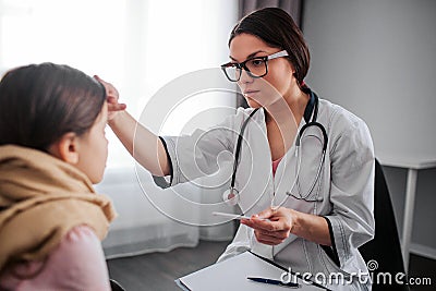 Worried young female doctor hold hand on child`s forehead. She look at her. Young woman hold thermometer in hand. They Stock Photo