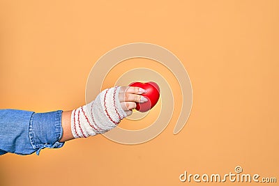 Wounded hand of caucasian young woman cover with bandage holding red heart over isolated yellow background Stock Photo