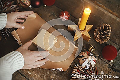 Wrapping christmas gift. Hands wrapping gift box in festive craft red paper on rustic wooden table with candle, scissors and Stock Photo