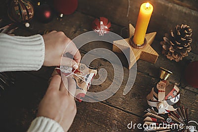 Wrapping christmas gift. Hands wrapping gift box in festive craft red paper on rustic wooden table with candle, scissors and Stock Photo