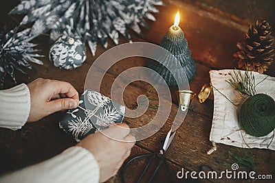 Wrapping christmas gift. Hands wrapping simple gift box in festive blue paper on rustic wooden table background with candle, Stock Photo