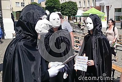 Three actors, dressed in white theater masks and black cloaks, entertain people at a festive fair on a city street. Editorial Stock Photo