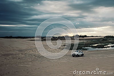 4x4 car waiting next to a lagoon in front of the sand dunes at sunset with beautiful sky Stock Photo