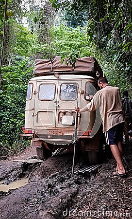 4x4 offroad vehicle stuck on muddy dirt road with young man trying to recover it in rain forest of Cameroon, Africa Editorial Stock Photo