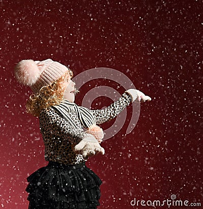 Xmas and New Year concept. Surprised baby girl in warm hat and gloves standing under snow, catching it on her open palms Stock Photo