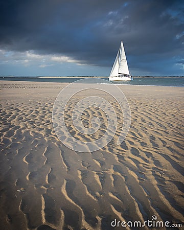 A yacht sailing with storm clouds over head and wet golden sands in the foreground Stock Photo