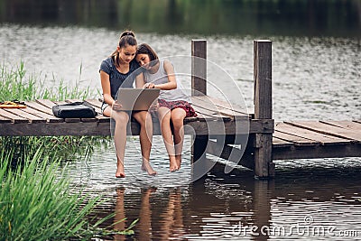 A 13-year-old sister and her 11-year-old sister sit Teach homework with a computer with an internet on a wooden bridge, The river Stock Photo