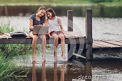 A 13-year-old sister and her 11-year-old sister sit Teach homework with a computer with an internet on a wooden bridge, The river Stock Photo