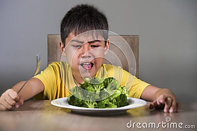 7 or 8 years old upset and disgusted Asian kid sitting on table in front of broccoli plate looking unhappy rejecting the fresh Stock Photo