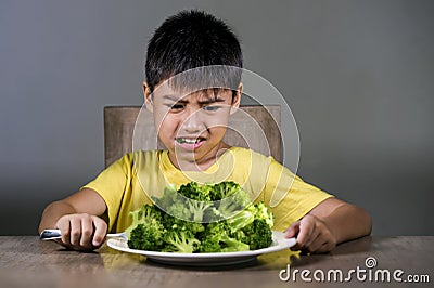 7 or 8 years old upset and disgusted Asian kid sitting on table in front of broccoli plate looking unhappy rejecting the fresh Stock Photo