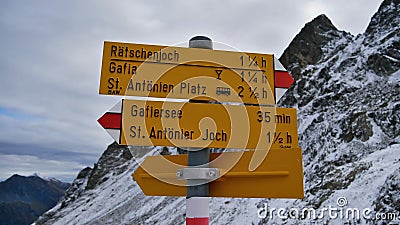 Yellow colored signpost showing hiking duration and directions to different destinations with snow-covered mountains. Stock Photo