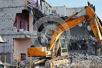 Yellow excavator demolishing a multi-storey building. Destroyed floors of the building, are pieces of stone, concrete, fittings. Stock Photo