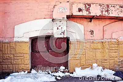 Yellow, pink colors wall with old red boarded up window on the ground level, ventilation pipe, melting snow on asphalt Stock Photo
