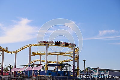 A yellow and red rollercoaster with people riding on the Santa Monica pier with lush green palms trees, blue sky and clouds Editorial Stock Photo