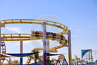 A yellow and red winding rollercoaster surrounded by colorful carnival rides with blue sky and light clouds at Santa Monica Pier Editorial Stock Photo