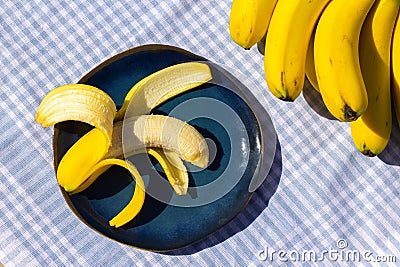 Yellow ripe bananas and one half peeled banana on the plate. Fresh organic fruits on blue tablecloth background on sunny day. Stock Photo