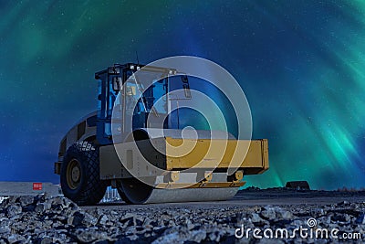 Yellow road roller stands on not ready new road under early night Aurora Borealis, left side view. Northern Lights shine over old Stock Photo