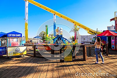 A yellow rollercoaster surrounded by colorful carnival rides and lush green palm trees on a brown wooden pier with clear blue sky Editorial Stock Photo